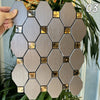 A hand holds a hexagonal mosaic tile pattern featuring silver and gold accents against a green plant background.