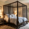 A large mesh fly net canopy is set up over a bed, featuring dark wood framing and sheer black netting.