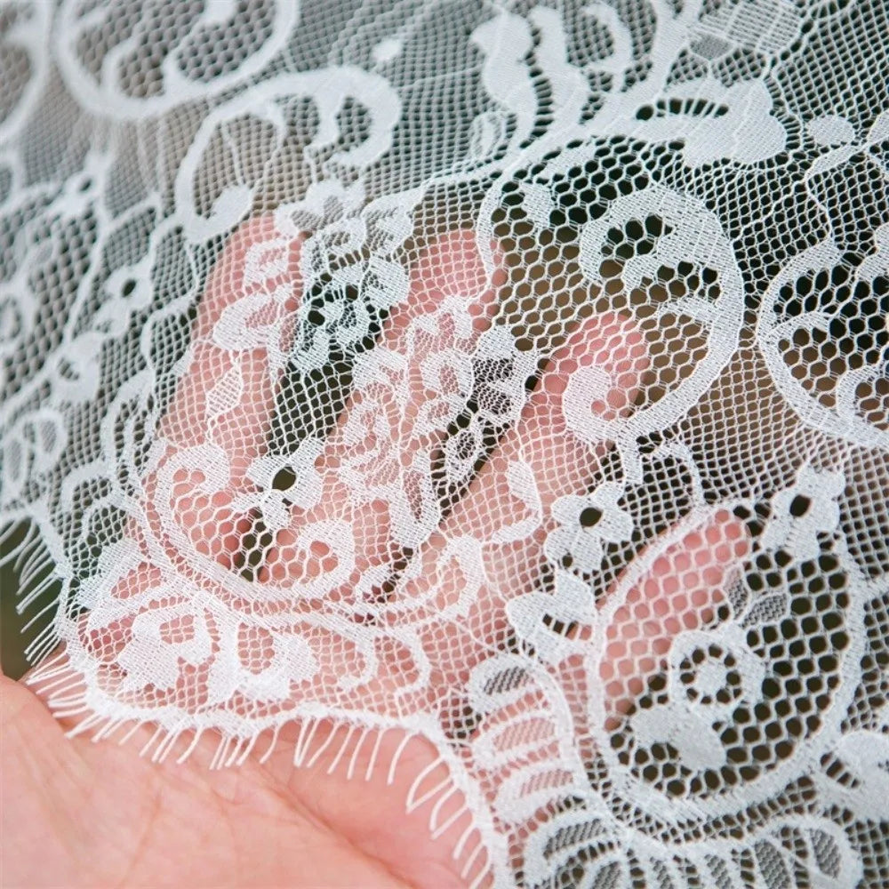 A hand is holding or touching white lace fabric featuring a French retro floral design.