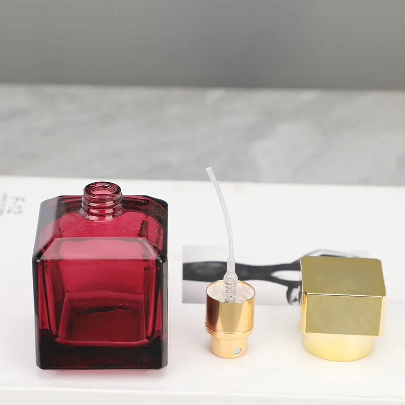 A red glass perfume bottle with a fine-mist sprayer and a gold-colored cap are displayed on a white surface.