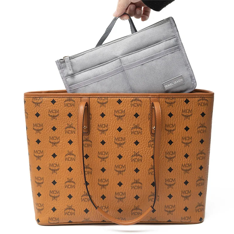A hand is placing a gray travel season purse organizer insert into a brown leather handbag.