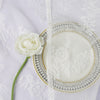 A white rose is placed next to a round, lace-edged fabric disc on a white cloth.