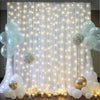 A backdrop featuring sheer white tulle curtains illuminated by warm string lights and surrounded by various clear and white b