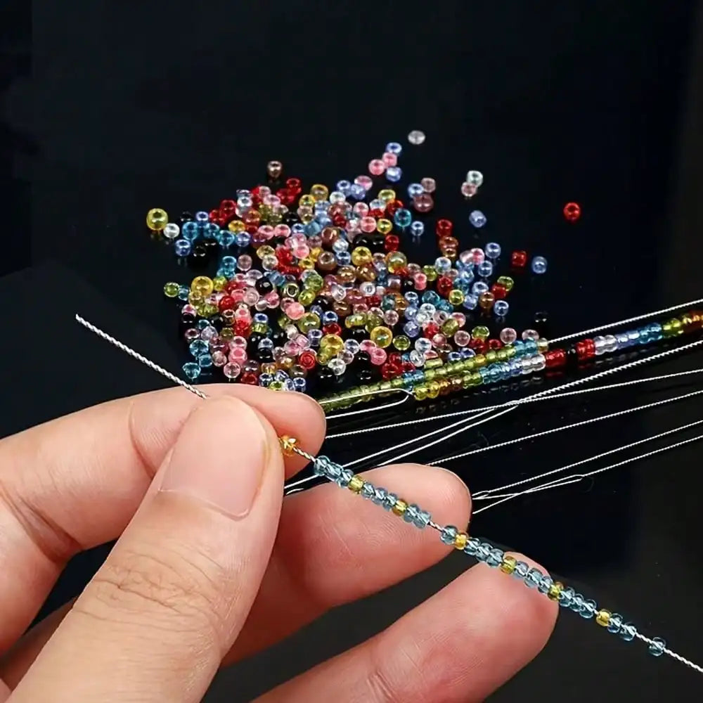 A hand holds a set of ultra-fine beading needles with various colored threads for jewelry making.