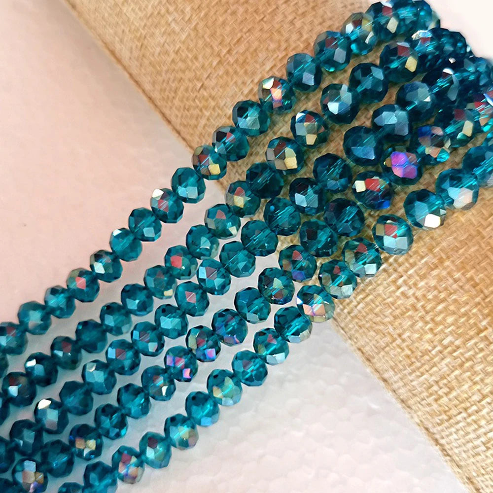 A strand of faceted blue-green crystal glass spacer beads is laid out on a beige fabric background.