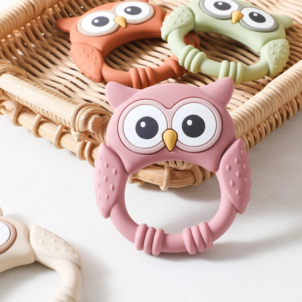 Three owl-shaped silicone teethers and rattles in orange, green, and pink resting in a woven basket.