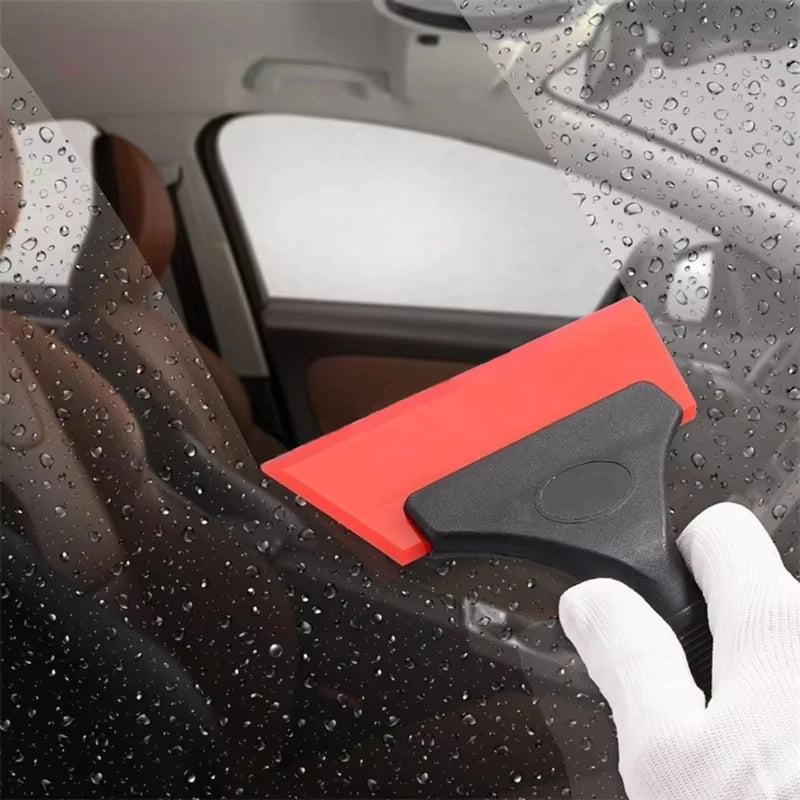 A hand uses an orange car window squeegee with a black handle to clean water droplets from a car window.