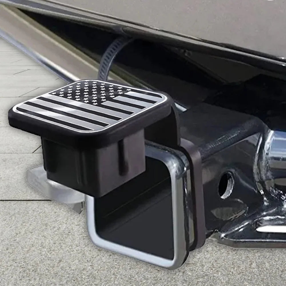 A black trailer hitch receiver cover with a flag emblem is shown attached to a vehicle's hitch.
