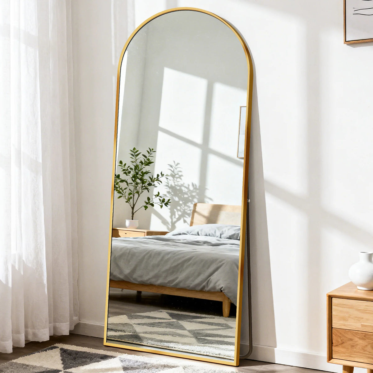 A full length oval mirror with a gold frame reflecting a bedroom and a window light.