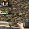 A hand is placing peel and stick waterproof tile stickers on a dark stone backsplash in a kitchen setting.