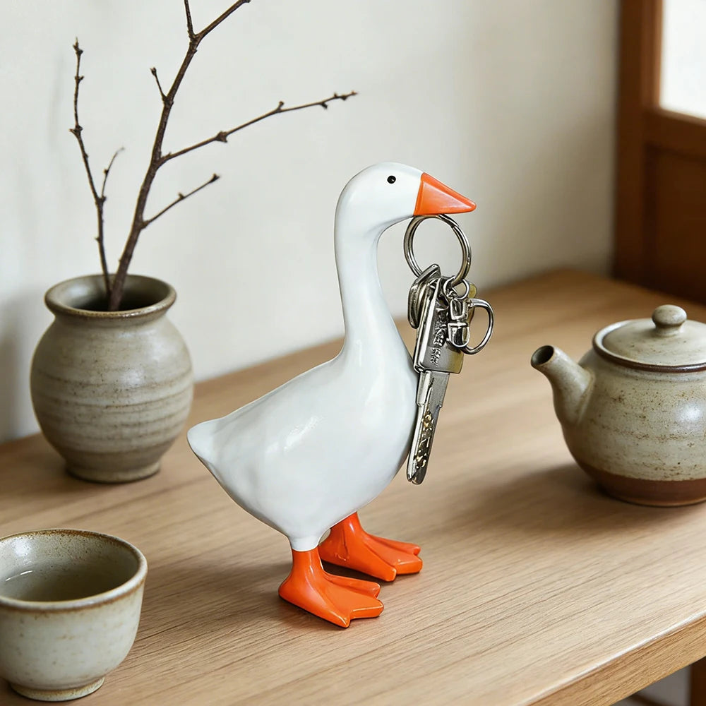 A white resin goose key holder sculpture with orange feet resting on a wooden desk.
