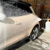 A silver car is being washed by a heavy-duty pressure washer, creating a foam spray on the body.