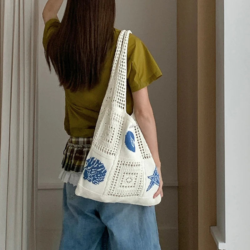 A person is holding a white handwoven knit tote bag with blue geometric patterns in a natural light setting.
