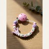 A pink silicone baby pacifier clip with a purple accent is displayed on a wooden surface.