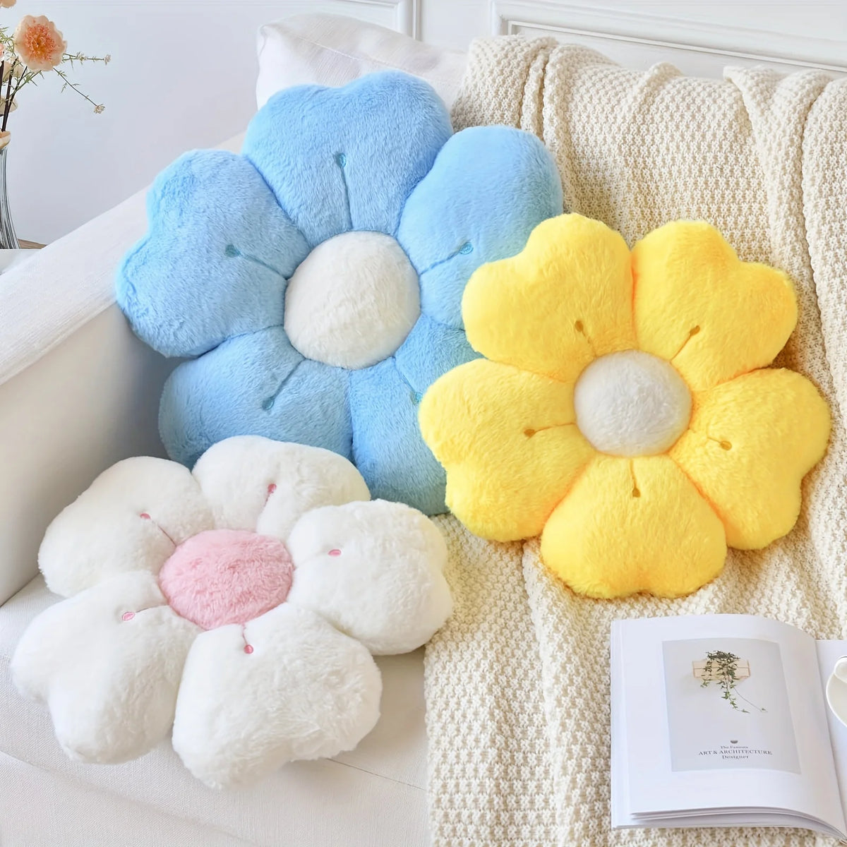 A collection of soft, fluffy daisy flower throw pillows in blue, yellow, and white resting on a cream-colored knitted blanket