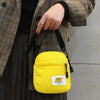 A yellow mini canvas crossbody bag is shown being held by a person wearing a plaid jacket and dark clothing.