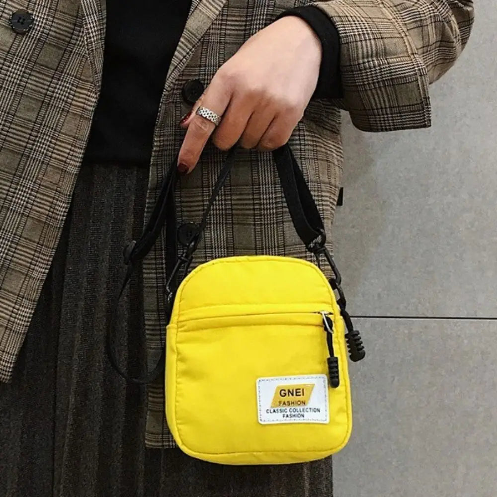 A yellow mini canvas crossbody bag is shown being held by a person wearing a plaid jacket and dark clothing.