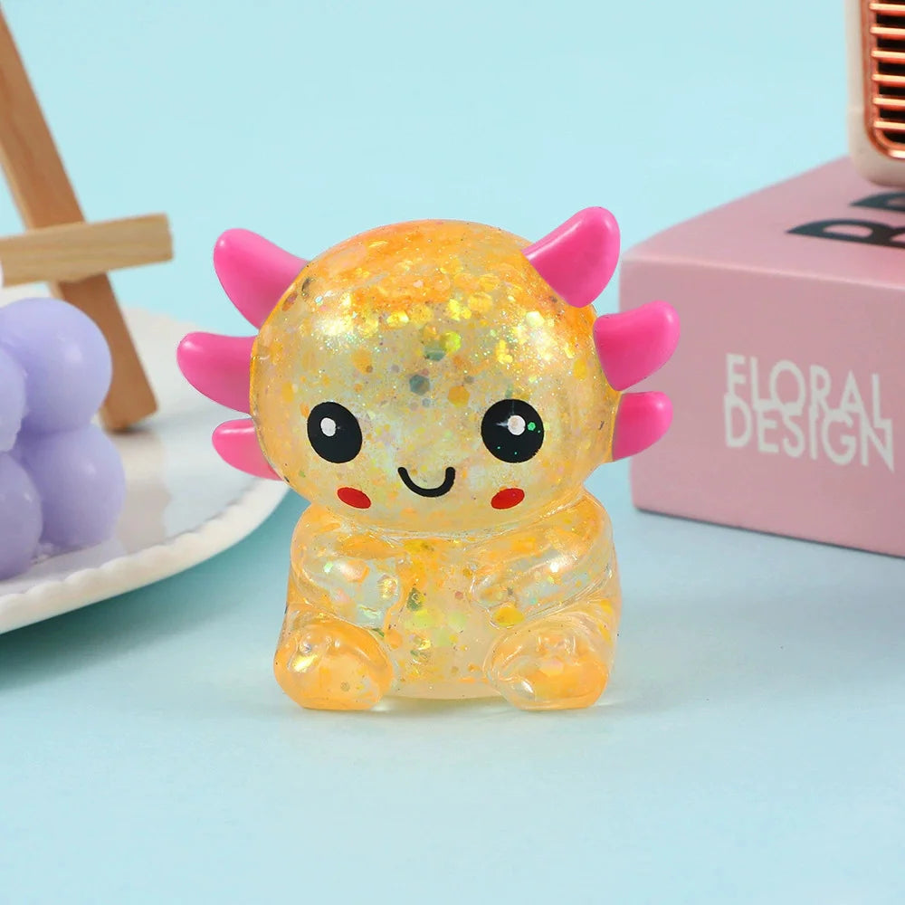 A small, squishy, translucent yellow axolotl stress ball with pink horns resting on a light blue surface.