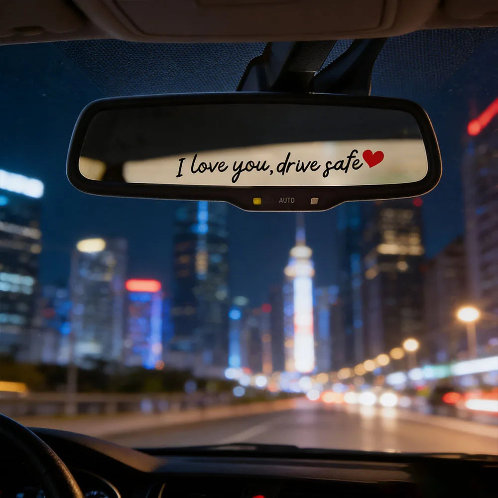 A car rearview mirror with a decal that reads I love you drive safe on a dark city street at night.