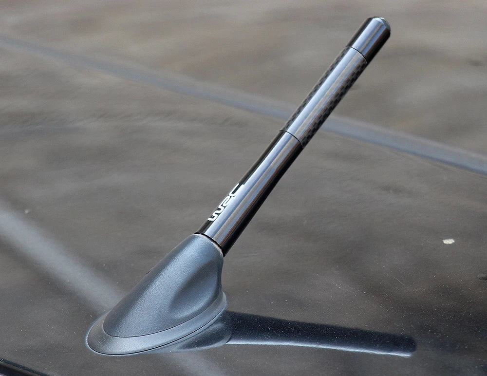 A black carbon fiber short radio antenna is shown angled against a dark surface.