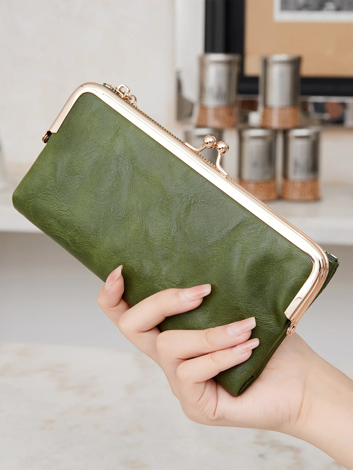 A hand holds a olive green leather wallet with gold accents against a blurred background.