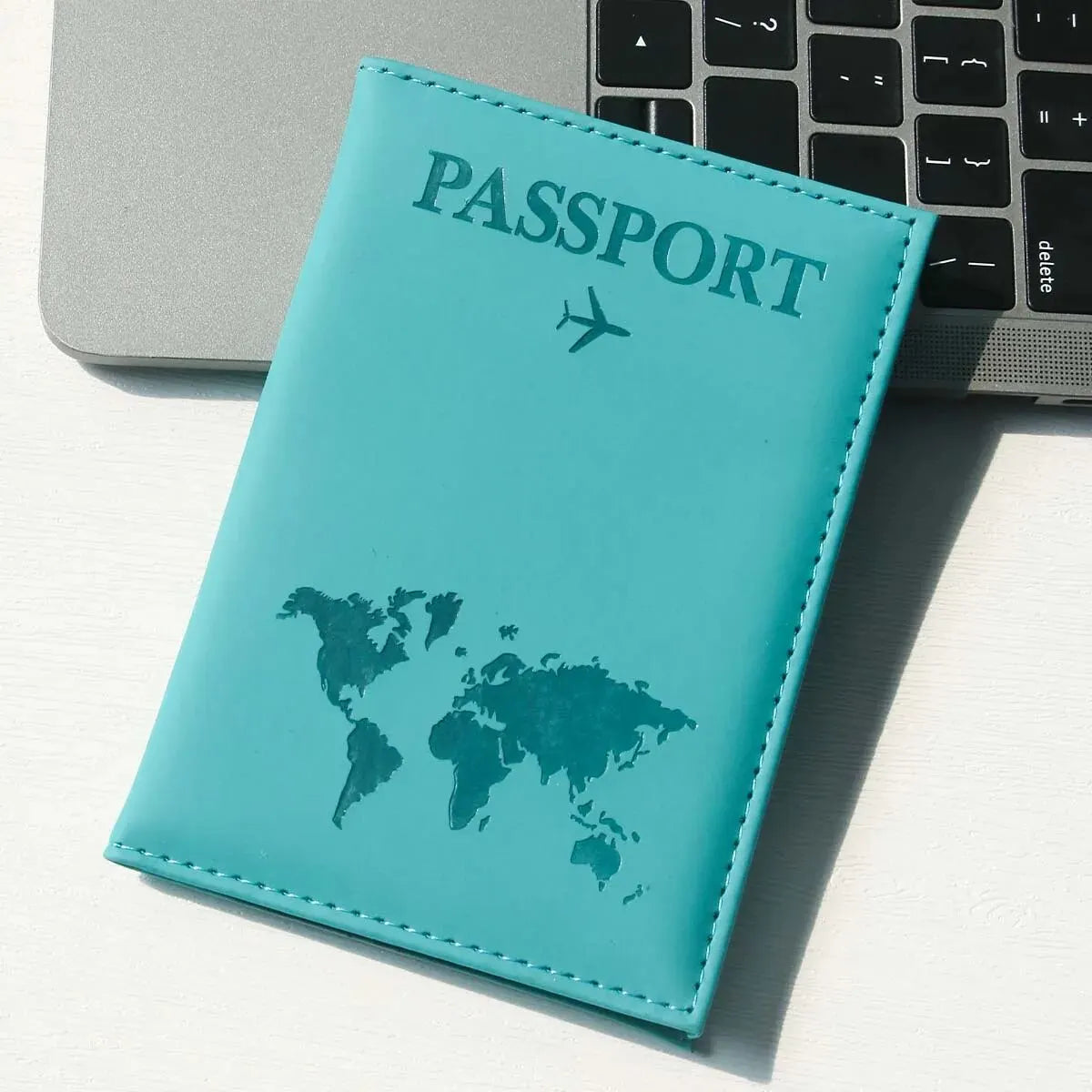 Türkis-Leder-Reisepasshülle mit Weltkartenmotiv auf weißem Untergrund neben einem Laptop.