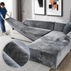 A person is draping a plush gray velvet elastic sofa cover over an L-shaped sofa.