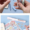 A hand holds a pink mini pen-shaped scissors and cutter tool next to various small plastic items.