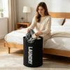 A woman sits on a bed and holds an extra-large black laundry basket with padded handles.