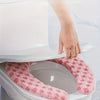 A hand is placing a plush pink winter toilet seat cushion cover onto a white toilet bowl.