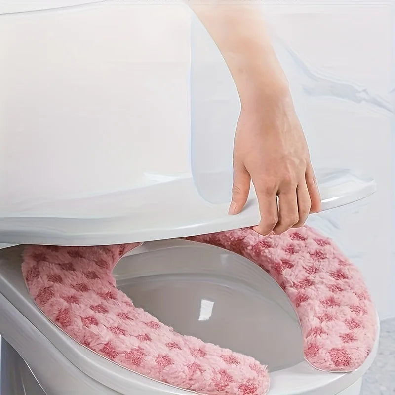 A hand is placing a plush pink winter toilet seat cushion cover onto a white toilet bowl.