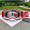 A pair of red and white motocross goggles resting on a white paper against a gray surface with greenery in the background.