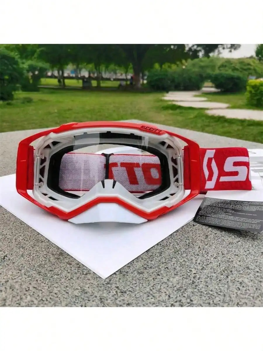 A pair of red and white motocross goggles resting on a white paper against a gray surface with greenery in the background.
