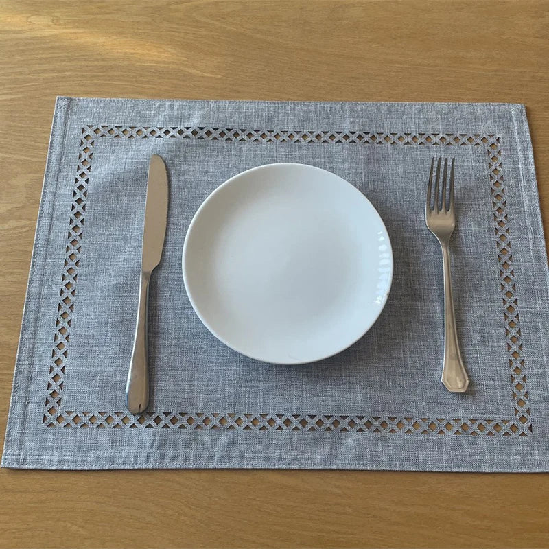 A place setting featuring a white plate, knife, and fork on a gray linen placemat with embroidered edges.