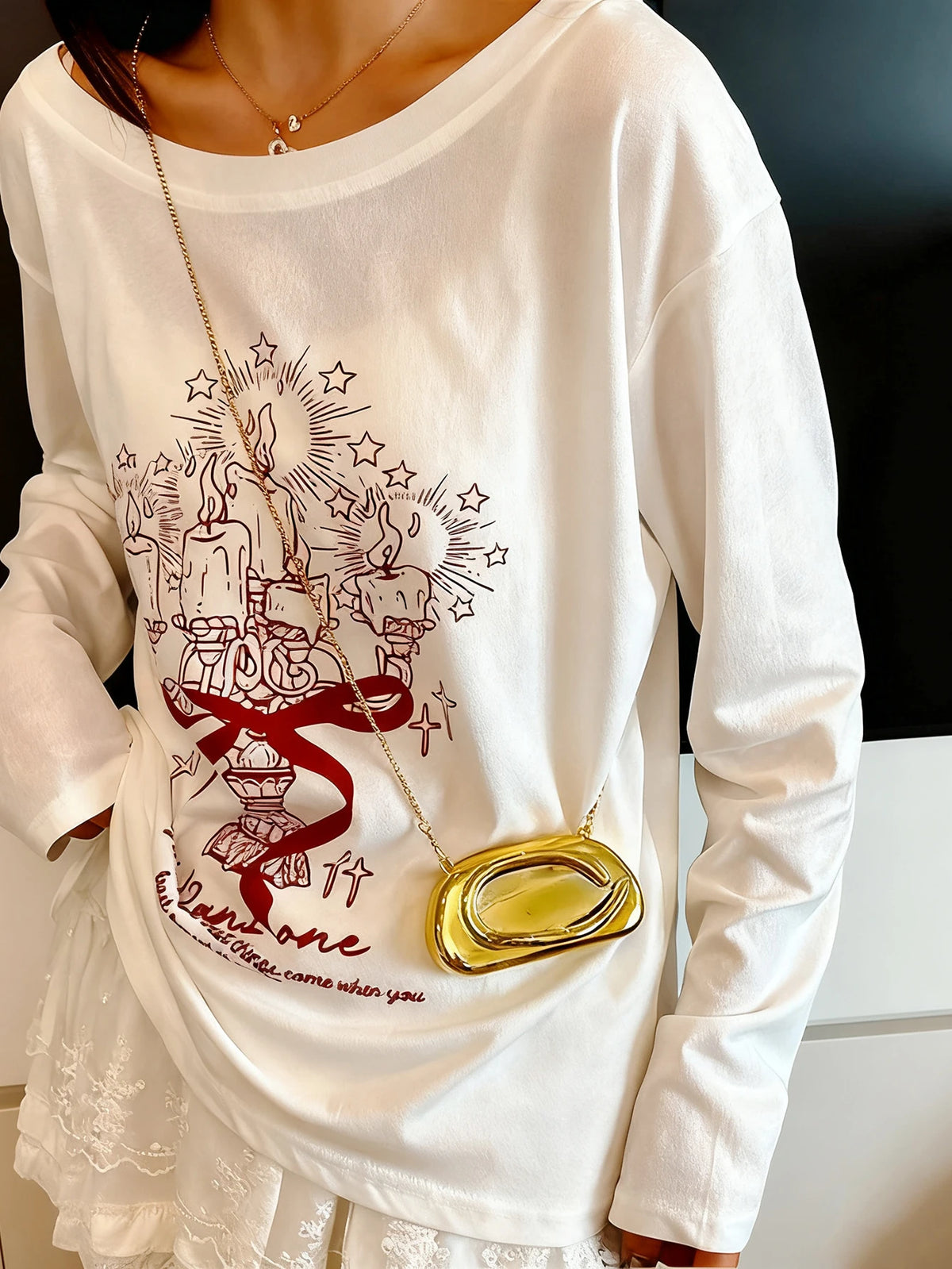 A person wearing a white long-sleeve shirt with a graphic design and a gold metal clutch bag.