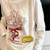 A person wearing a white long-sleeve shirt with a graphic print is holding a mini gold metal clutch bag.