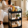 A woman is using a black 3-tier rolling utility cart with wheels to store various items on a light rug.