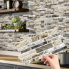 A hand is placing peel-and-stick tile stickers onto a kitchen backsplash made of stacked marble tiles.