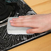 A hand is placing a white cleaning sponge onto a black non-stick surface for kitchen cleaning.