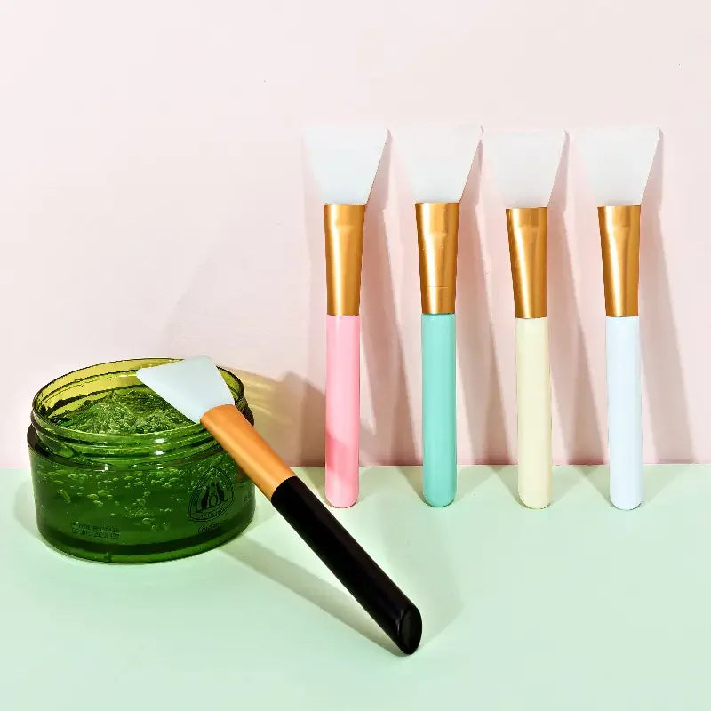 A collection of silicone facial mask brushes with gold and colored handles displayed on a light surface.