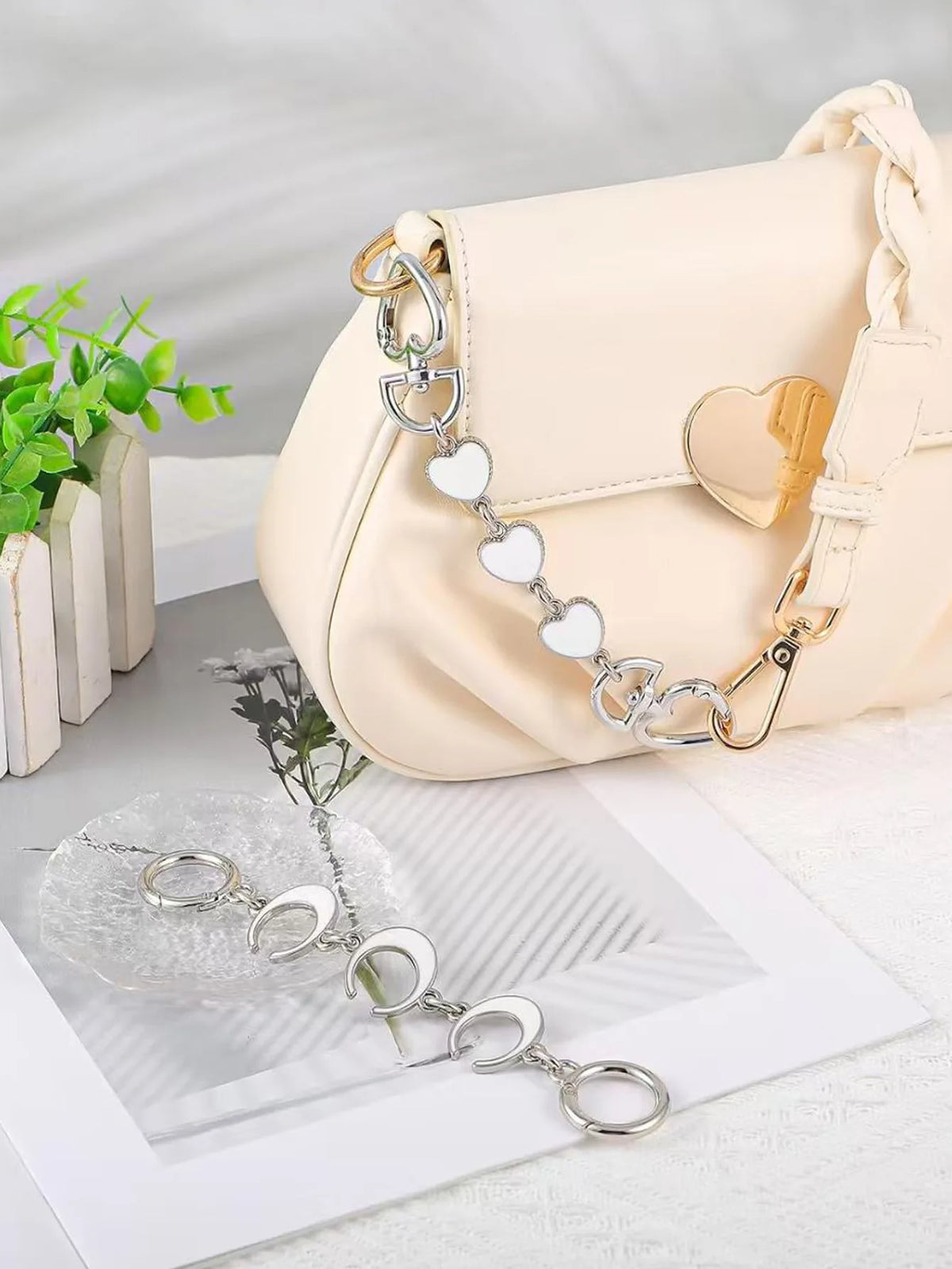 Helle beige Lederhandtasche mit goldener Kettenerweiterung und silbernen Herzanhängern auf weißem Untergrund.