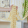 A hand is holding a bohemian tassel curtain made of light yellow or cream-colored fabric with numerous hanging lanterns.