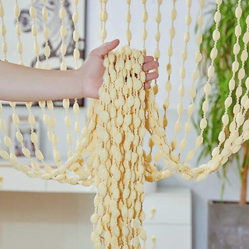 A hand is holding a bohemian tassel curtain made of light yellow or cream-colored fabric with numerous hanging lanterns.