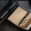 A close-up of a black leather refillable journal lying next to a light brown paper folder and a silver pen on a dark wooden s
