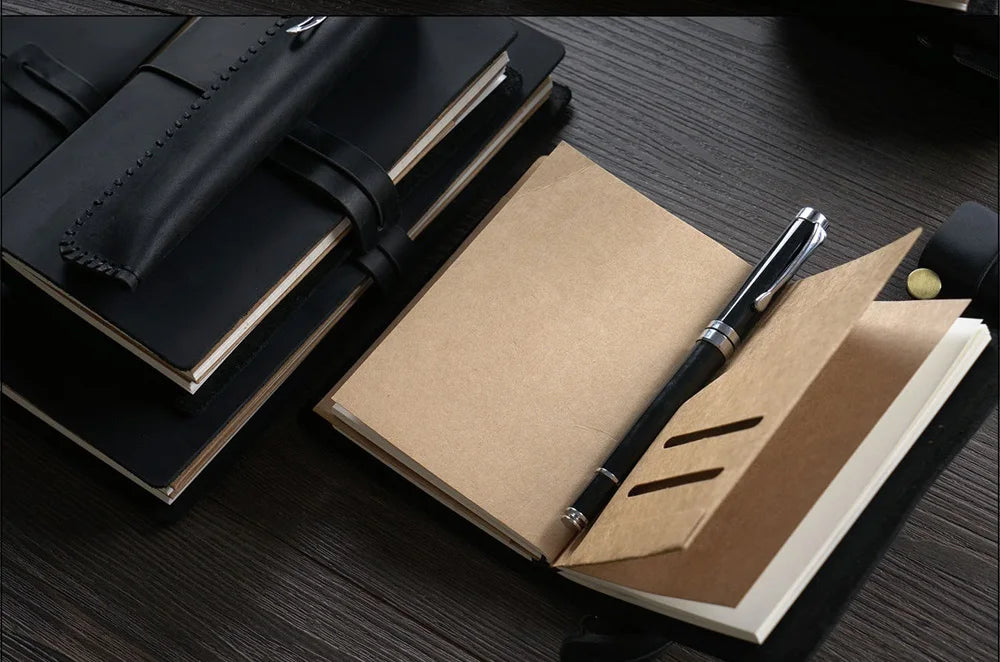 A close-up of a black leather refillable journal lying next to a light brown paper folder and a silver pen on a dark wooden s