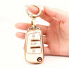 A hand holds a gold-toned key fob cover with an oval shape and a circular ring, featuring several buttons.