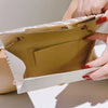 A hand holds an open pleated glitter envelope clutch in a beige color with a gold chain strap.