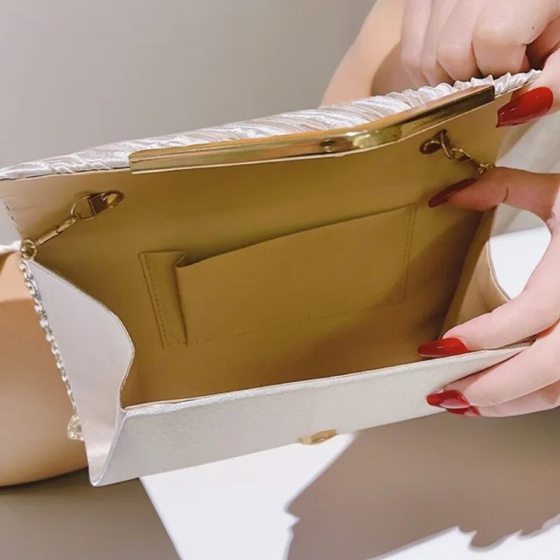 A hand holds an open pleated glitter envelope clutch in a beige color with a gold chain strap.