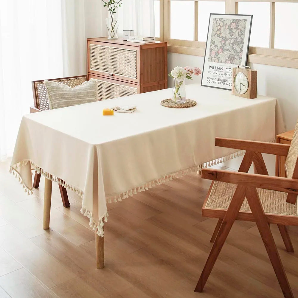 A rectangular dining table covered with a cream tablecloth featuring delicate cotton tassels, set in a bright room.