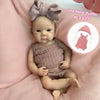 A realistic reborn silicone doll wearing a pink knitted outfit and a matching bow, lying on pink fabric.